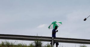 A man walks along a highway barrier under the scorching sun, shielding himself with a green and white umbrella during a heatwave.Photo credit: Carbon Brief
