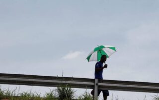A man walks along a highway barrier under the scorching sun, shielding himself with a green and white umbrella during a heatwave. Photo credit: Carbon Brief